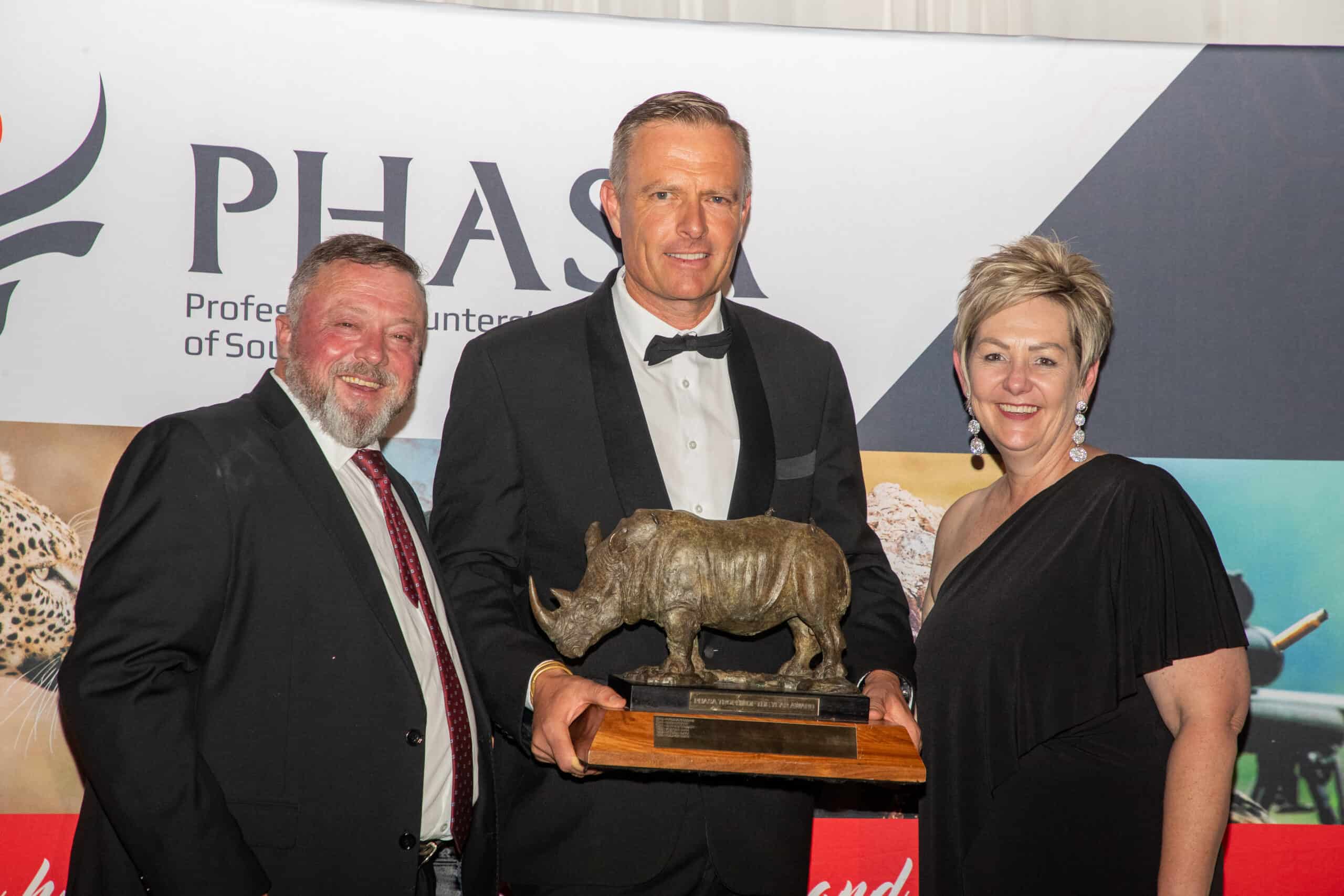 Three people at awards event, one holding a rhino trophy, in front of Graham Sales Safaris banner.